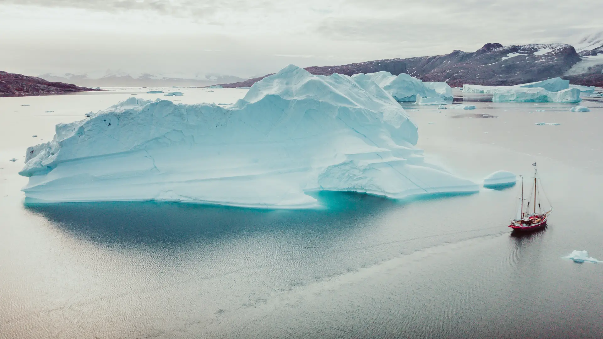 A red sailboat passing a vast turquoise iceberg in a calm northern fjord